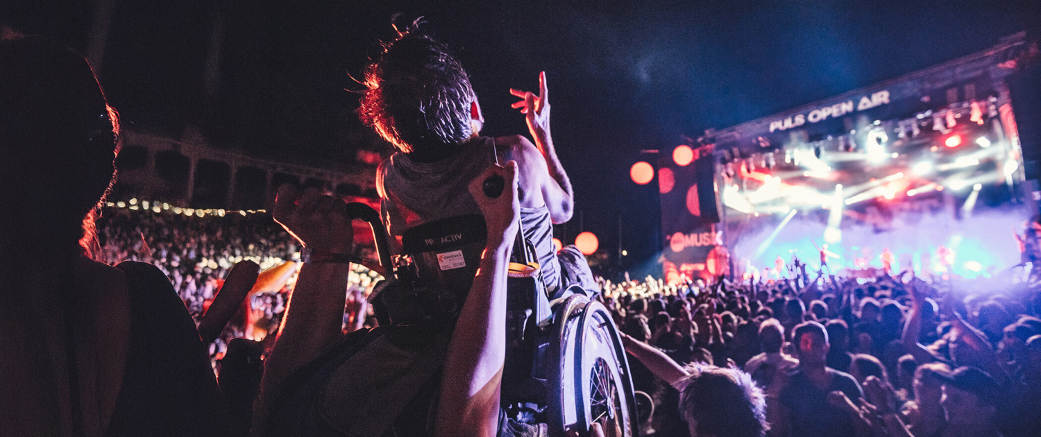 Eine Person im Rollstuhl wird bei einem Musikfestival von der Menge hochgehoben. Vor einer großen, bunt beleuchteten Bühne feiern viele Menschen bei Nacht.