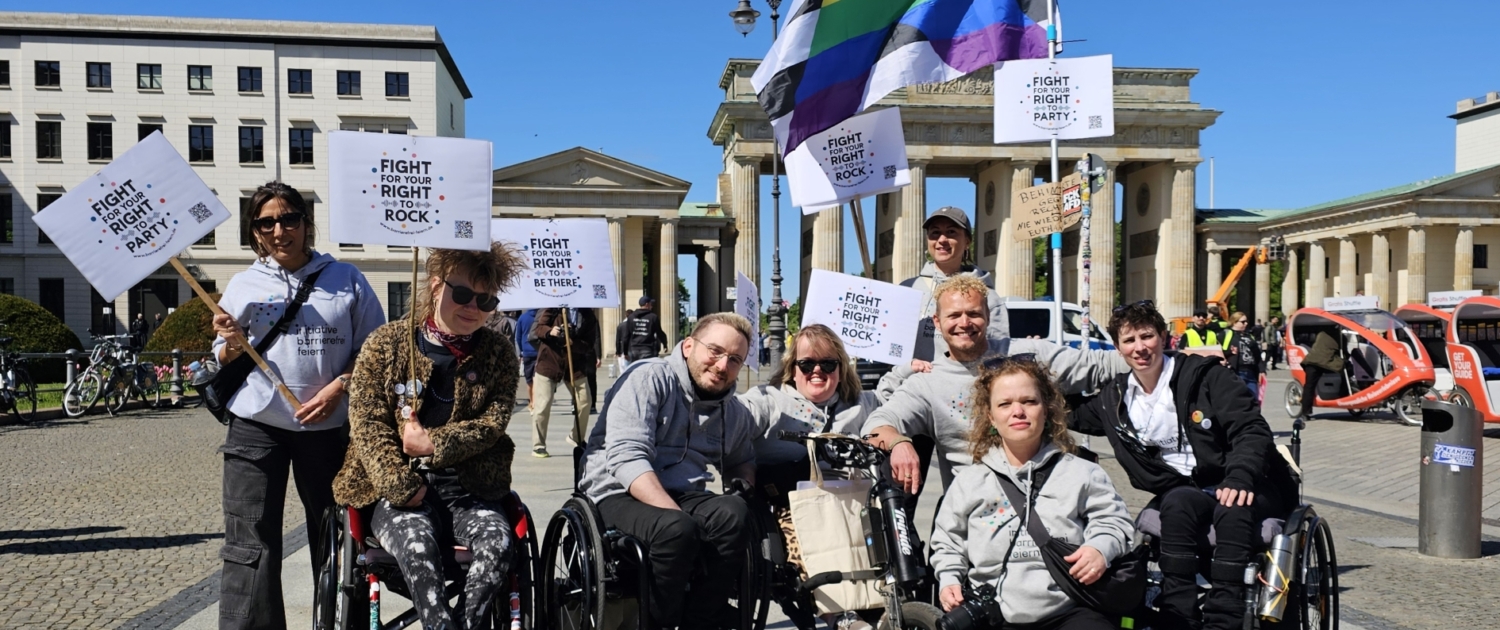 Acht Personen mit und ohne Behinderung mit Demonstrations-Schildern und einer Disability Flagge vor dem Brandenburger Tor. Auf den Schildern steht: "Fight For Your Right To Party!"